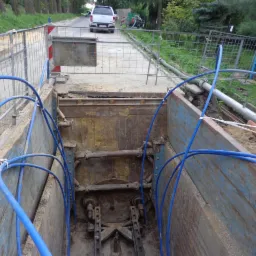 Wykop zabezpieczony metalowymi szalunkami podczas budowy przyłącza wodno-kanalizacyjnego; widoczne niebieskie rury, fragment drogi i przejeżdżający samochód.