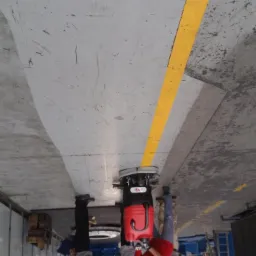 Przemysłowa maszyna czyszcząca z czerwonym zbiornikiem, usuwająca zabrudzenia z betonowej posadzki w hali magazynowej, widoczne ślady opon i żółta linia wyznaczająca strefę.