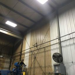 Centro-Tech Karol Dybowski - Pracownik w kasku, na podnośniku, czyści metalową ścianę hali przemysłowej. Część ściany jest już umyta, widać różnicę w kolorze. Na ścianie widoczne rury i wentylator.