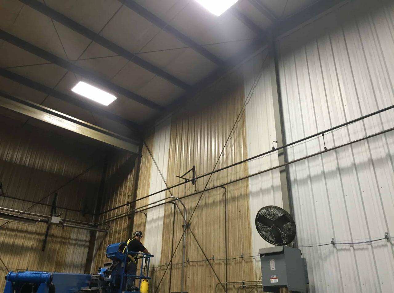 Pracownik w kasku, na podnośniku, czyści metalową ścianę hali przemysłowej. Część ściany jest już umyta, widać różnicę w kolorze. Na ścianie widoczne rury i wentylator.