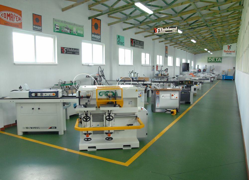 Wnętrze hali produkcyjnej z maszynami do obróbki drewna, widoczne różne modele i marki, w tym Tecnomax i Robland, pod sufitem reklamy producentów maszyn.