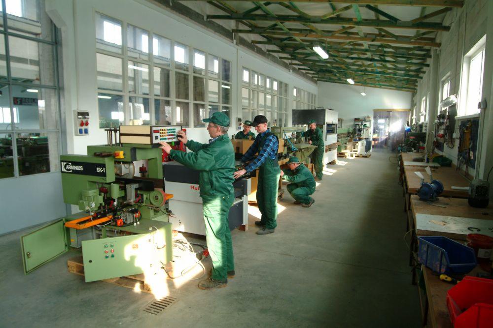 Pracownicy w zielonych kombinezonach obsługują maszyny do obróbki drewna w przestronnej hali produkcyjnej z dużymi oknami i drewnianą konstrukcją stropu.