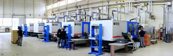 Wnętrze hali produkcyjnej z rzędem wtryskarek, widoczni pracownicy obsługujący maszyny, system rur pod sufitem.