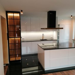 Max Fornir Design - Kuchnie na wymiar - Wałbrzych - Nowoczesna kuchnia z wyspą i czarnym blatem, oświetleniem LED pod szafkami i przeszkloną szafką z drewnianym wnętrzem. Kontrast czerni i bieli, połyskujące powierzchnie.