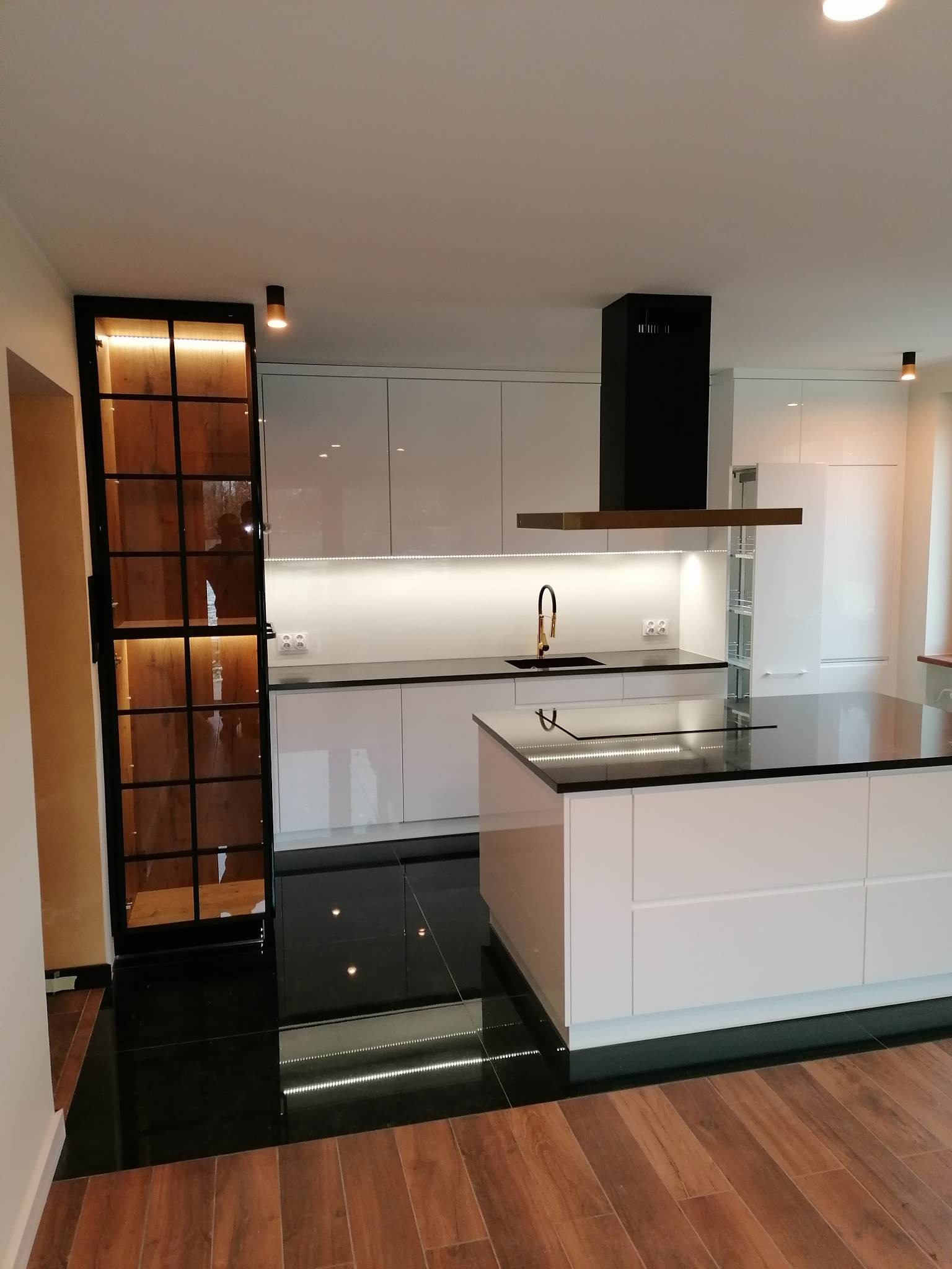 Nowoczesna kuchnia z wyspą i czarnym blatem, oświetleniem LED pod szafkami i przeszkloną szafką z drewnianym wnętrzem. Kontrast czerni i bieli, połyskujące powierzchnie.
