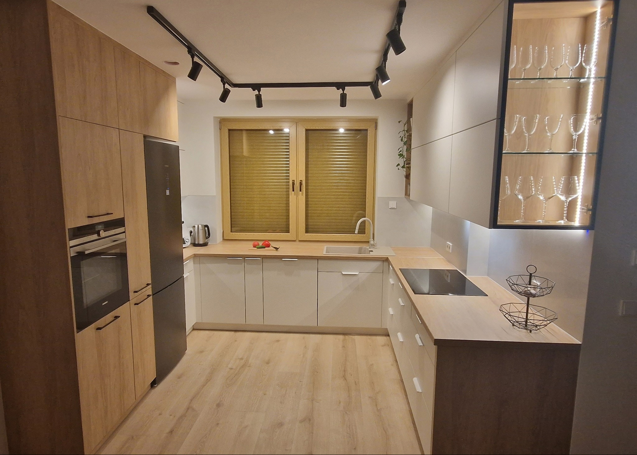 Jasna kuchnia na wymiar z drewnianymi akcentami, oświetleniem punktowym i przeszkloną szafką z kieliszkami. Nowoczesny design z elementami naturalnego drewna.