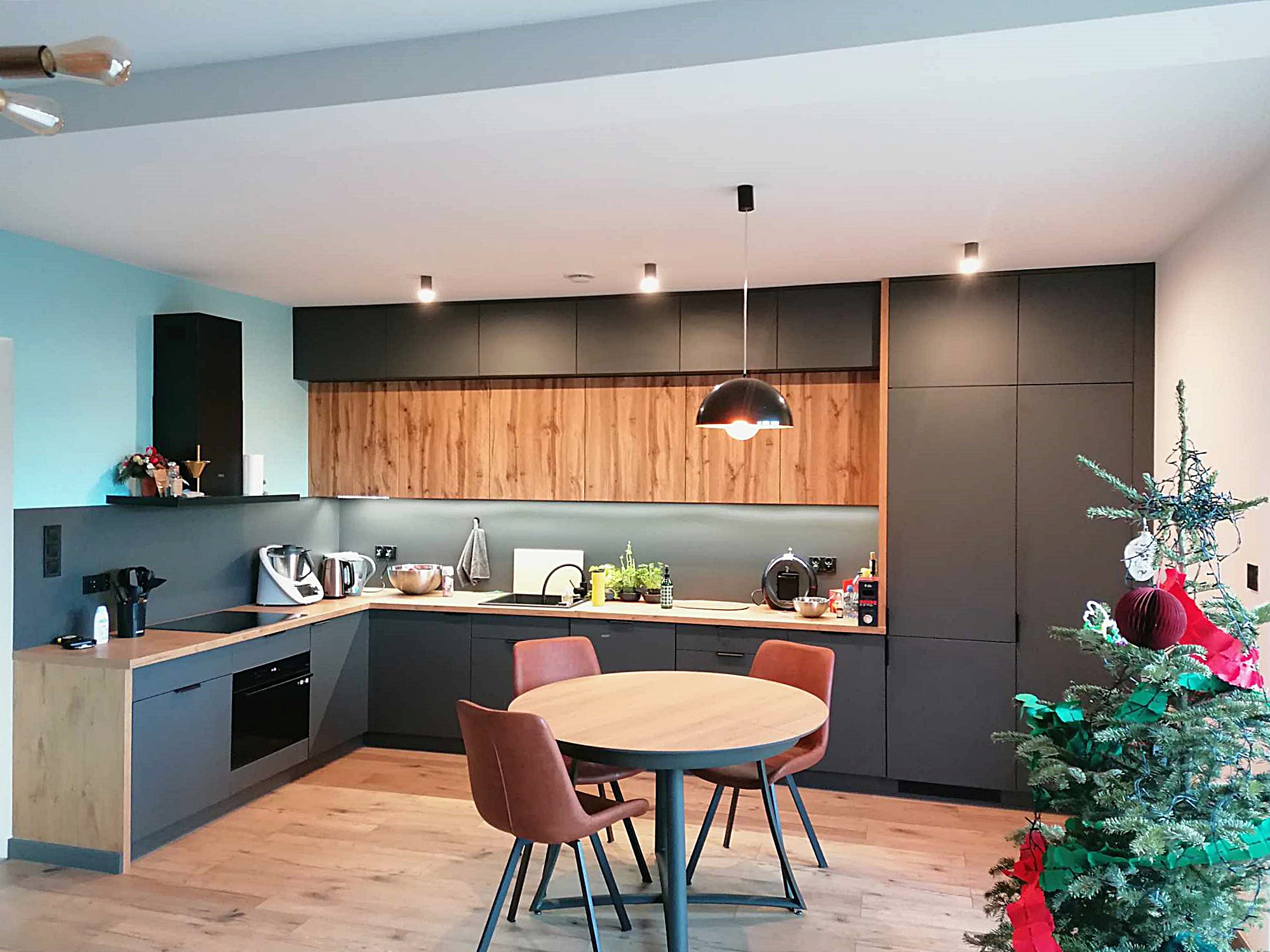 Nowoczesna kuchnia z ciemnymi szafkami i drewnianym panelem nad blatem. Okrągły stół z krzesłami i choinka w rogu. Oświetlenie punktowe i wisząca lampa.