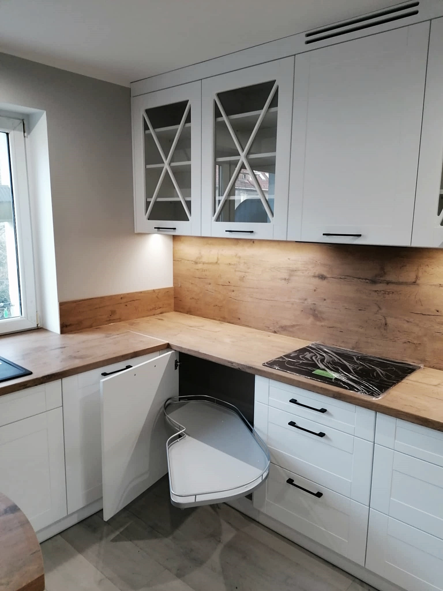 Jasna kuchnia na wymiar z drewnianym blatem i szafkami z przeszklonymi frontami. Otwarta szafka narożna z obrotowym systemem przechowywania.