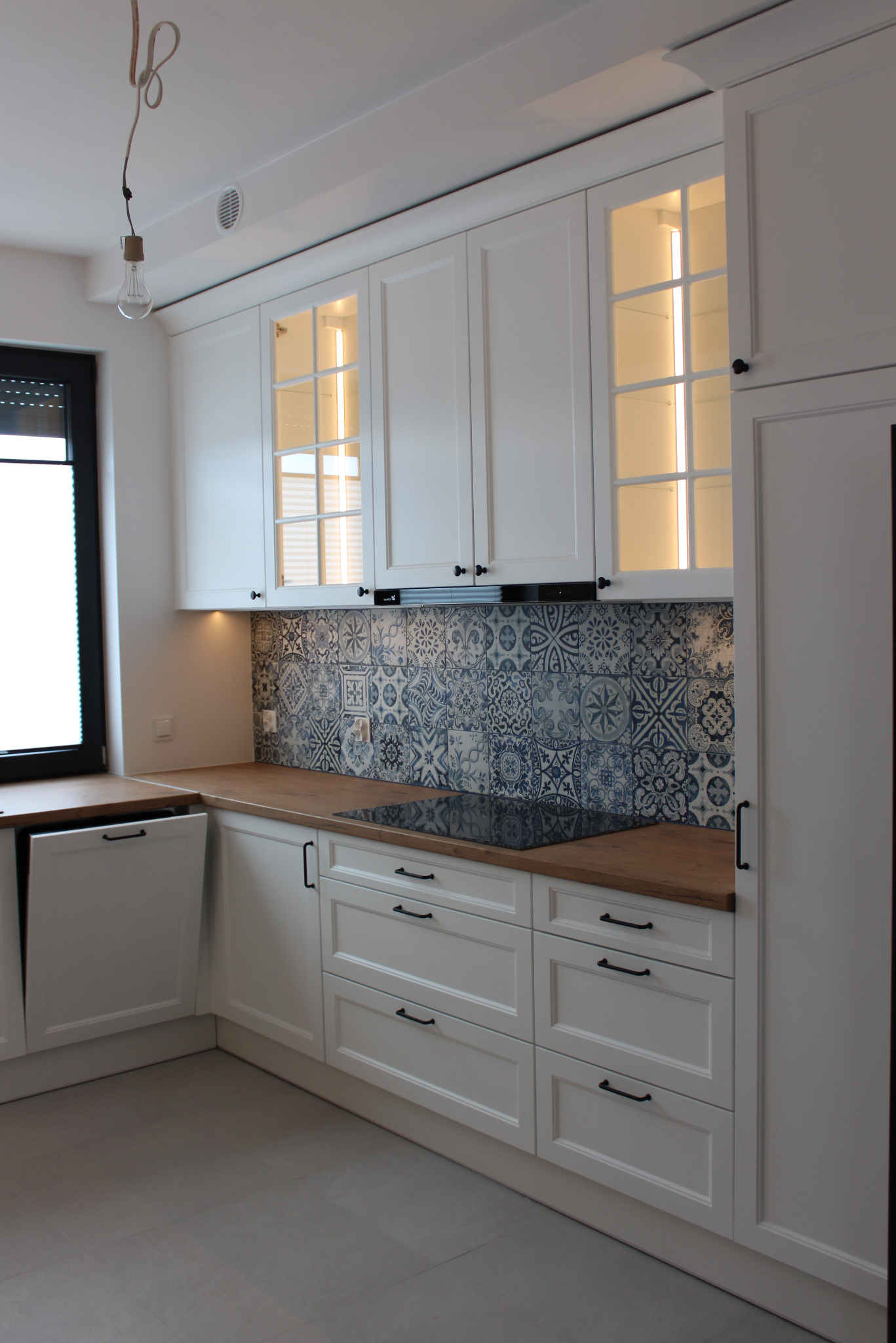 Biała kuchnia na wymiar z drewnianym blatem i dekoracyjnymi płytkami nad indukcją. Szafki górne z przeszkleniami podświetlone od wewnątrz. Minimalistyczne czarne uchwyty.