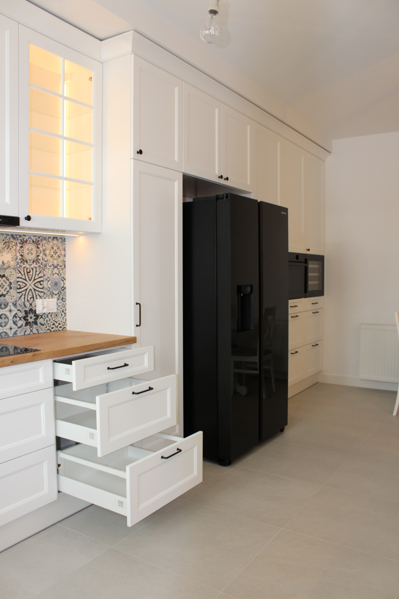 Biała kuchnia na wymiar z otwartymi szufladami, drewnianym blatem i czarną lodówką. Dekoracyjne płytki nad blatem. Nowoczesne oświetlenie podszafkowe.