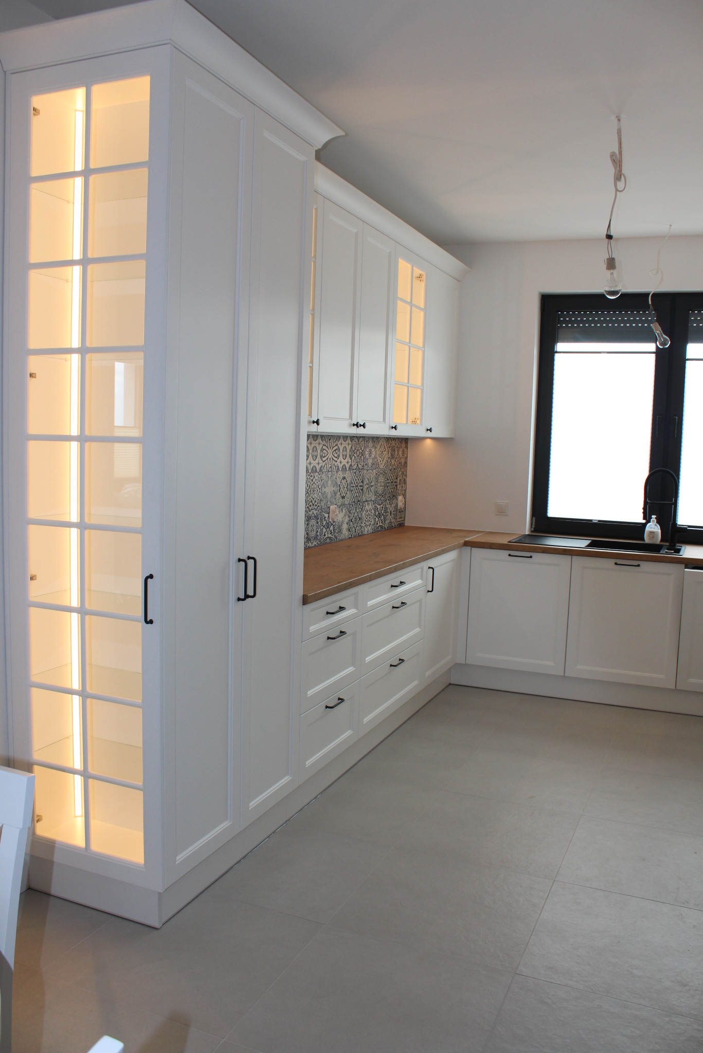 Jasna kuchnia z białymi szafkami, drewnianym blatem i czarnymi uchwytami. Szafka z podświetlanymi, przeszklonymi drzwiami. Nowoczesny design, płytki nad blatem.