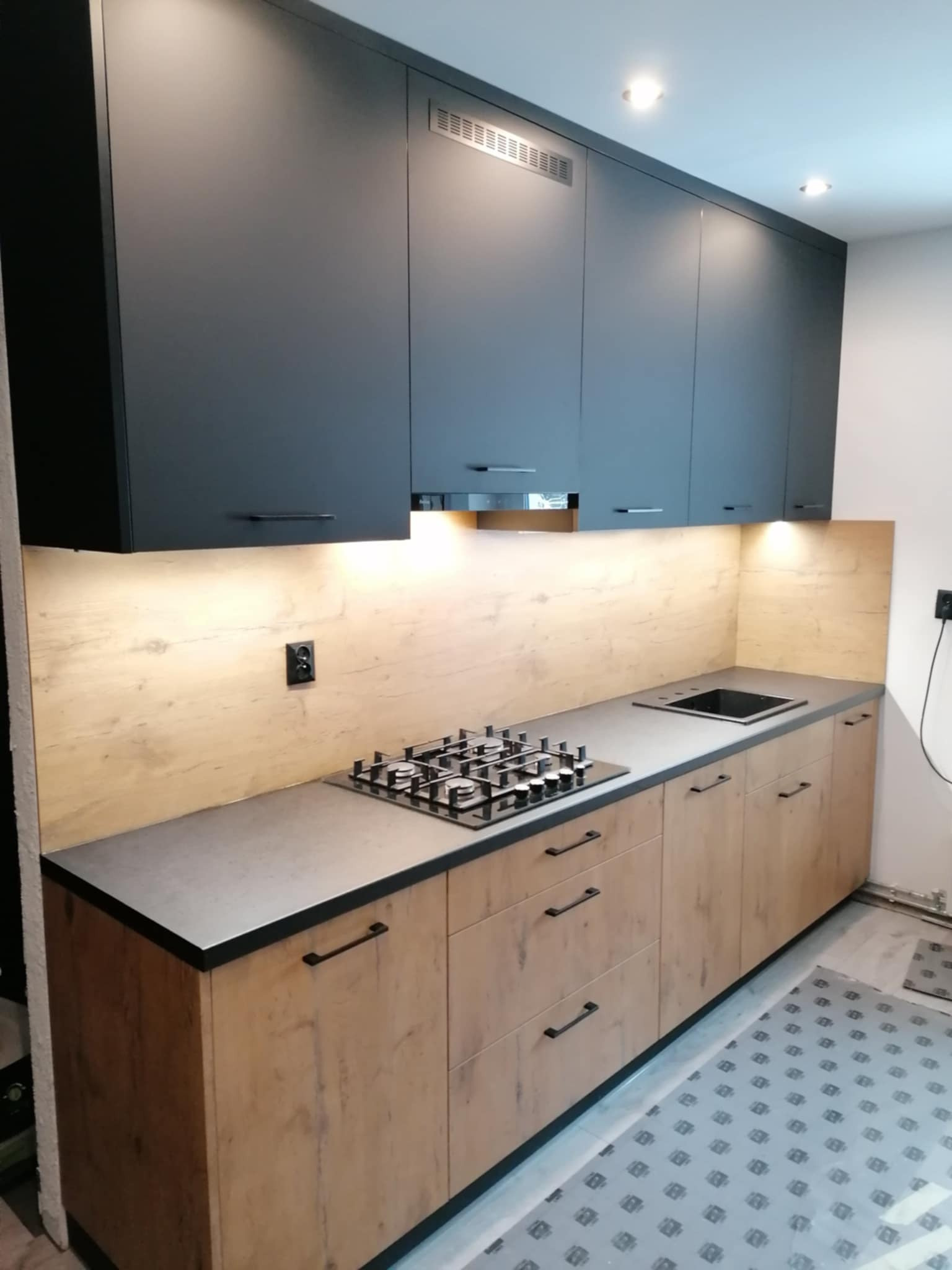 Nowoczesna kuchnia na wymiar z szarymi szafkami górnymi i drewnopodobnymi dolnymi, oświetlona listwą LED pod szafkami. Widoczna płyta gazowa i szary blat.