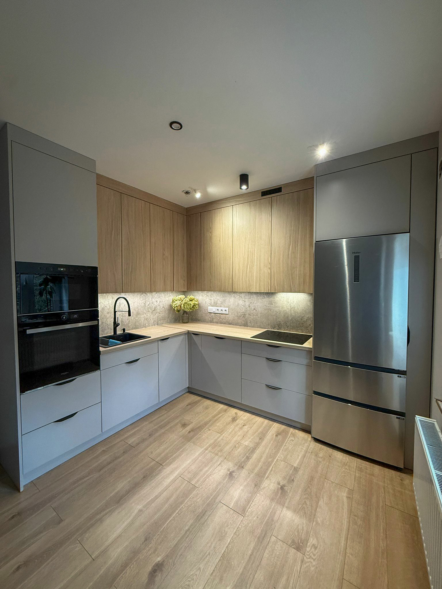 Jasna kuchnia na wymiar z drewnianymi i szarymi frontami, czarną armaturą i sprzętem AGD ze stali nierdzewnej. Uwagę przykuwają minimalistyczne lampy sufitowe.