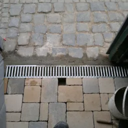 Dwa rodzaje nawierzchni: szara kostka granitowa przechodzi w beżowo-szarą kostkę brukową. Pomiędzy nimi zamontowany jest metalowy odpływ liniowy. Widoczne wiadro i kielnia.