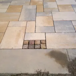 Ułożona kostka brukowa w odcieniach beżu i szarości z wstawką z kostki granitowej, widoczny piasek i mokre plamy.