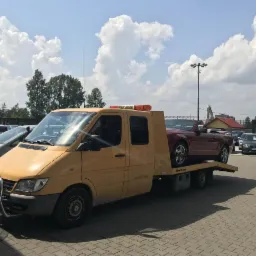 Żółta laweta Mercedes-Benz Sprinter z pomarańczowym światłem ostrzegawczym przewozi czerwony kabriolet na parkingu z innymi samochodami w tle, widoczne chmury na niebie.