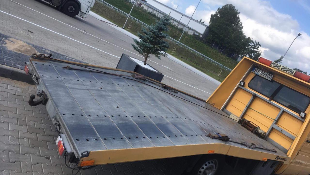 Żółta laweta z napisem 'Pomoc Drogowa' oraz numerem rejestracyjnym WS 80600, stojąca na parkingu z kostki brukowej, na platformie widoczna mała choinka w donicy.