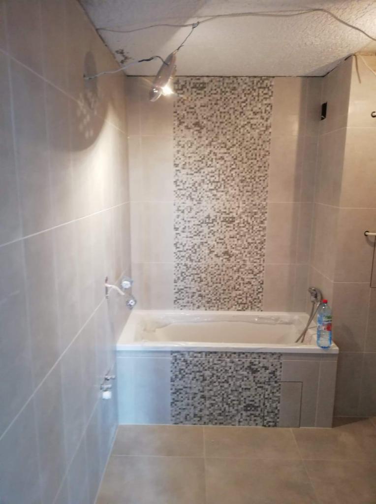 Łazienka w trakcie remontu: świeżo położone, beżowe płytki ścienne i podłogowe, z dekoracyjnym pasem mozaiki nad wanną, sufit w stanie surowym z wystającymi kablami.