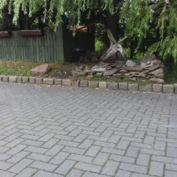 Szara kostka brukowa ułożona w jodełkę, obrzeże z kostki granitowej, w tle drewniana budka i ozdobna konstrukcja z kamieni i gałęzi pod drzewem.