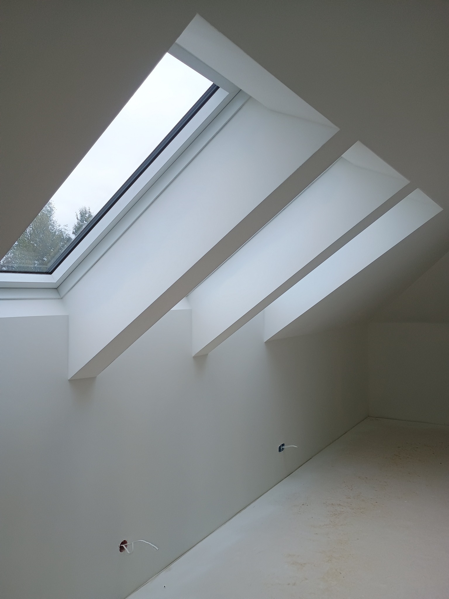 Białe poddasze z oknem dachowym i skośnymi belkami, widoczne kable elektryczne wystające ze ściany, minimalistyczny design, surowe wykończenie.