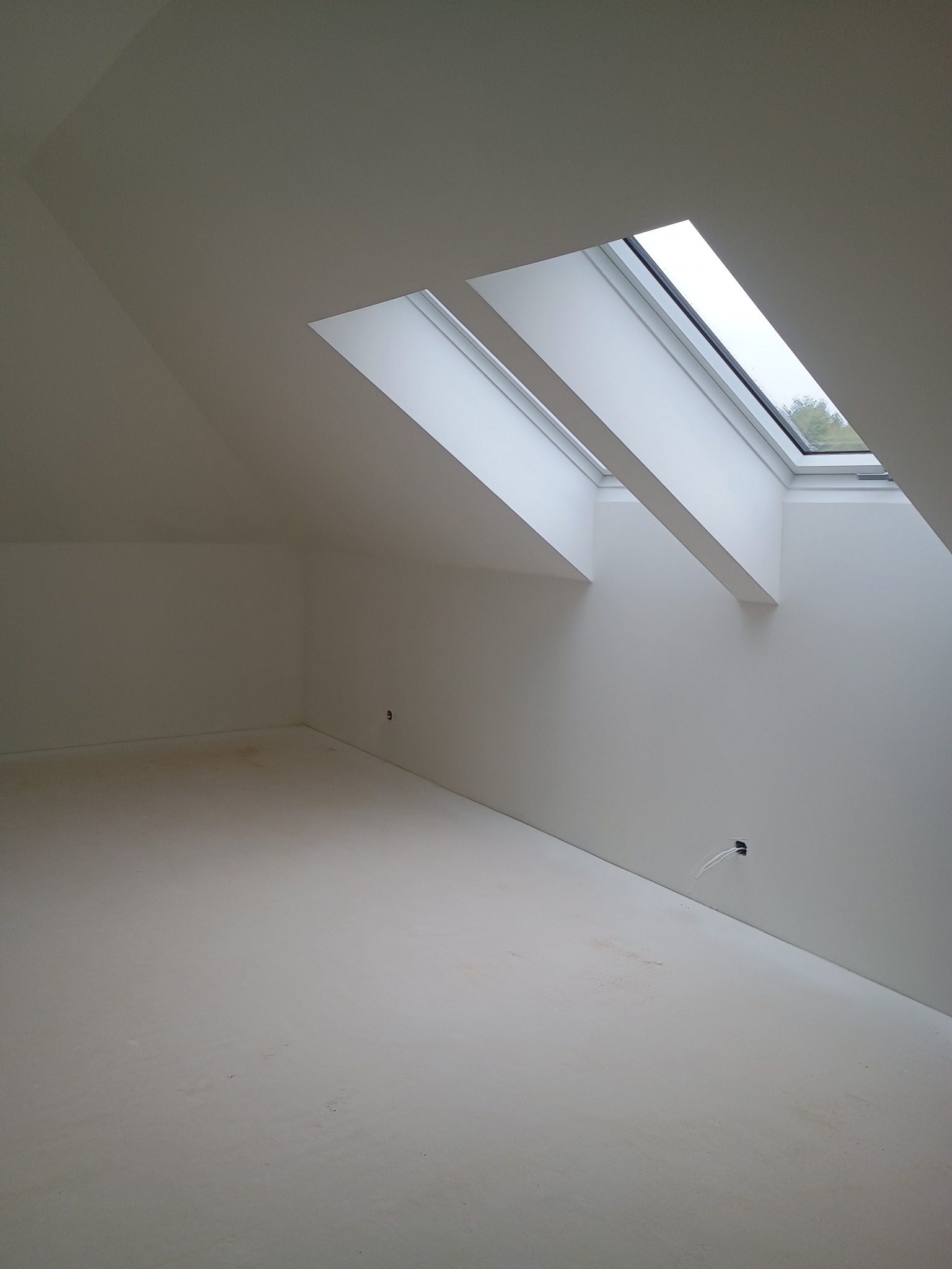 Jasne poddasze z dwoma oknami dachowymi, świeżo pomalowane na biało. Widoczne gniazdko elektryczne i listwa przypodłogowa. Minimalistyczny design, neutralne oświetlenie.