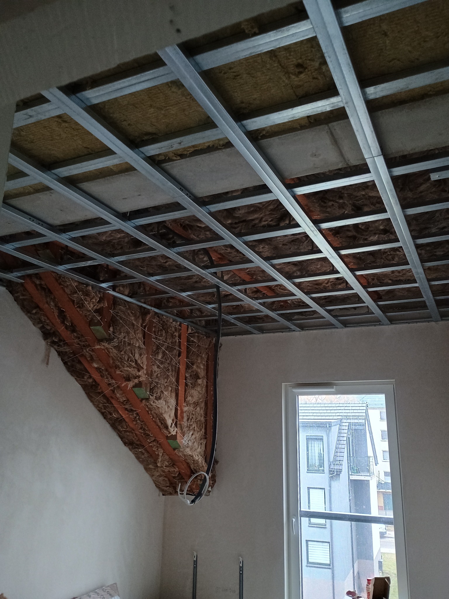 Sufit podwieszany w trakcie montażu: metalowa konstrukcja, izolacja z wełny mineralnej, częściowo zamontowane płyty. Widoczne okno i fragment ściany.