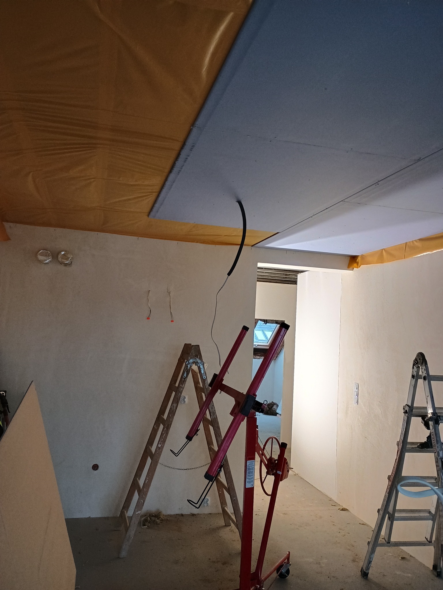 Montaż płyty gipsowej na suficie z użyciem podnośnika. Widoczne kable, drabiny i folia ochronna. Prace wykończeniowe w trakcie realizacji.