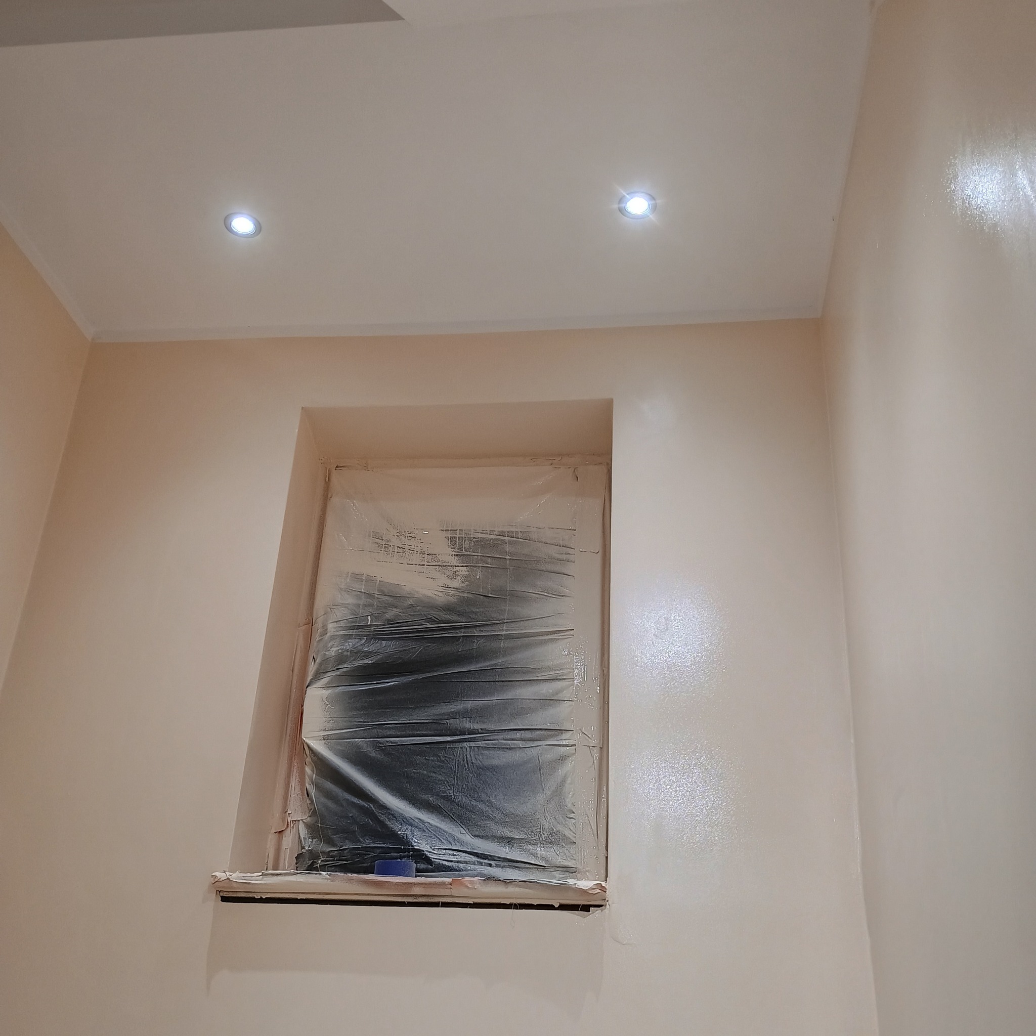 Świeżo pomalowane ściany w odcieniach beżu, okno zabezpieczone folią malarską, widoczne dwa punkty świetlne w suficie. Gładka powierzchnia ścian.