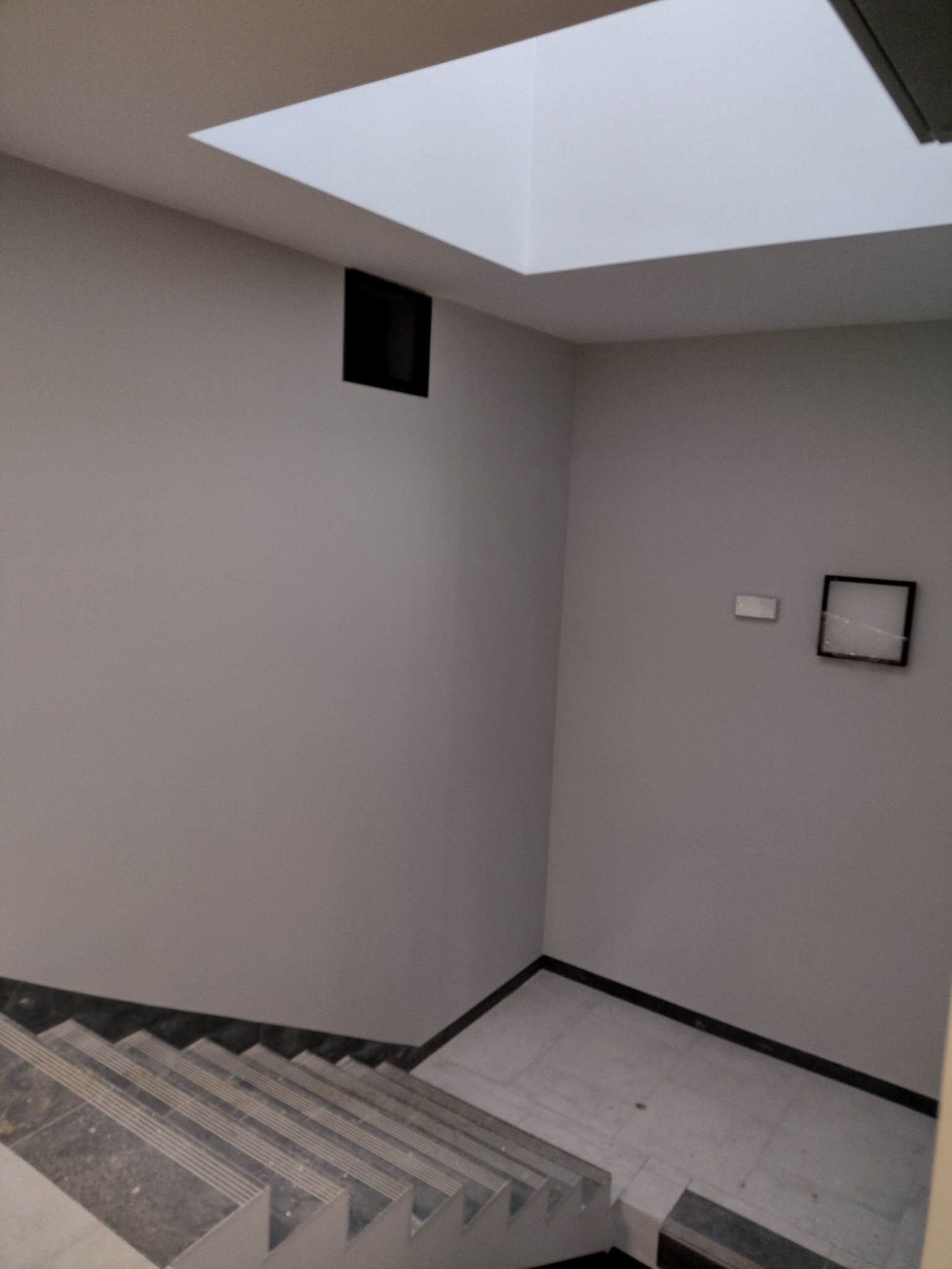 Szare ściany klatki schodowej z marmurowymi schodami i kwadratowym oknem dachowym. Minimalistyczne wykończenie wnętrza z czarnymi akcentami.