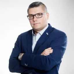 Portret mężczyzny w okularach i niebieskiej marynarce z rękami skrzyżowanymi na piersiach, na białym tle.