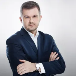 Portret mężczyzny w granatowym garniturze i białej koszuli, z założonymi rękami i srebrnym zegarkiem na nadgarstku, na jasnoszarym tle.