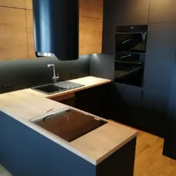 Nowoczesna kuchnia w ciemnej kolorystyce z drewnianymi akcentami, widoczny okap nad płytą grzewczą i zlewozmywakiem, zabudowane piekarniki.