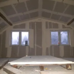 Poddasze w trakcie wykańczania: widoczne płyty na stelażu, przygotowane do dalszych prac, dwa okna z widokiem na zewnątrz, drewniane belki stropowe.
