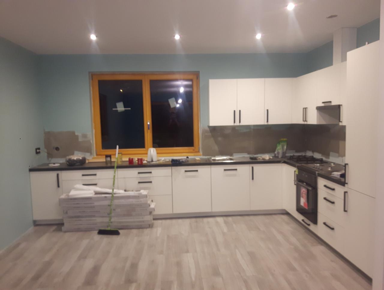 Nowoczesna, biała kuchnia z czarnymi akcentami w trakcie montażu, widoczne płyty na podłodze i ślady po pracach na ścianie.