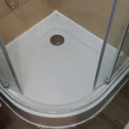 Narożna kabina prysznicowa z brodzikiem, widok z góry, z widocznymi elementami instalacji wodnej i odpływowej, oraz fragmentem umywalki i pralki, na podłodze imitacja drewnianych paneli.