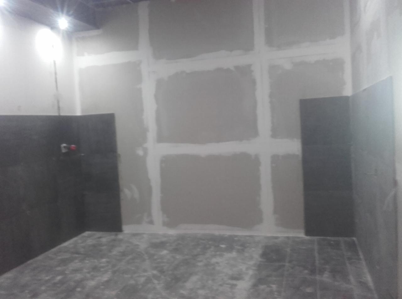 Wnętrze łazienki w trakcie prac: ściany przygotowane pod gładzie z widocznymi łączeniami płyt kartonowo-gipsowych, częściowo ułożone ciemne płytki na ścianie i podłodze, widoczne przyłącza wodne.