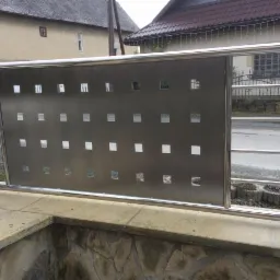 Nowoczesna balustrada ze stali nierdzewnej z perforowanym panelem i kwadratowymi otworami, zamontowana na kamiennym murku przed budynkiem.