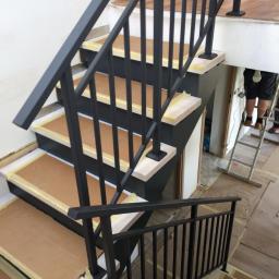 SZM Sp. z o.o. - Montaż czarnej, metalowej balustrady schodowej z pionowymi prętami, widoczne zabezpieczenia schodów kartonem i taśmą malarską, drabina w tle.