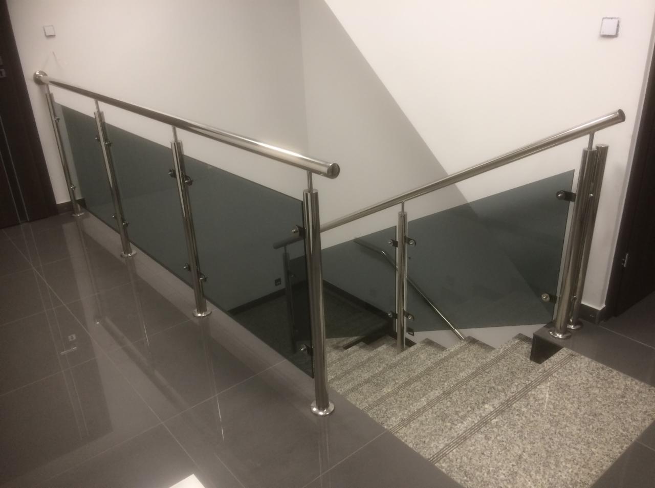 Wewnętrzna klatka schodowa z szarymi, szklanymi panelami i metalowymi poręczami, widoczna z góry, z szarymi płytkami na podłodze i schodach.