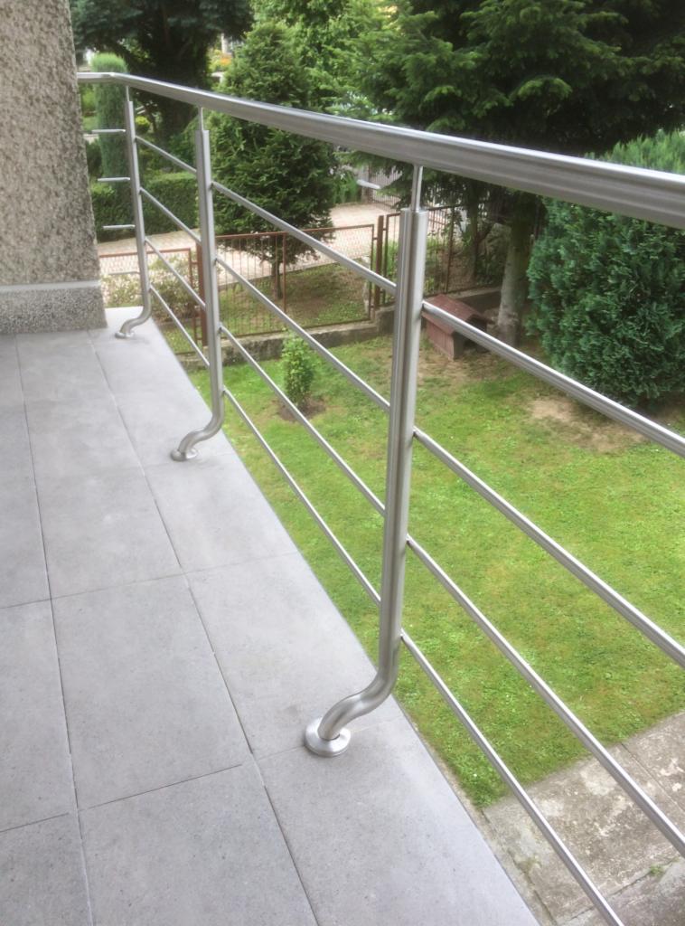 Nowoczesna balustrada nierdzewna z poziomymi prętami na balkonie z szarymi płytkami, widok na zadbany ogród z zielonym trawnikiem i krzewami.