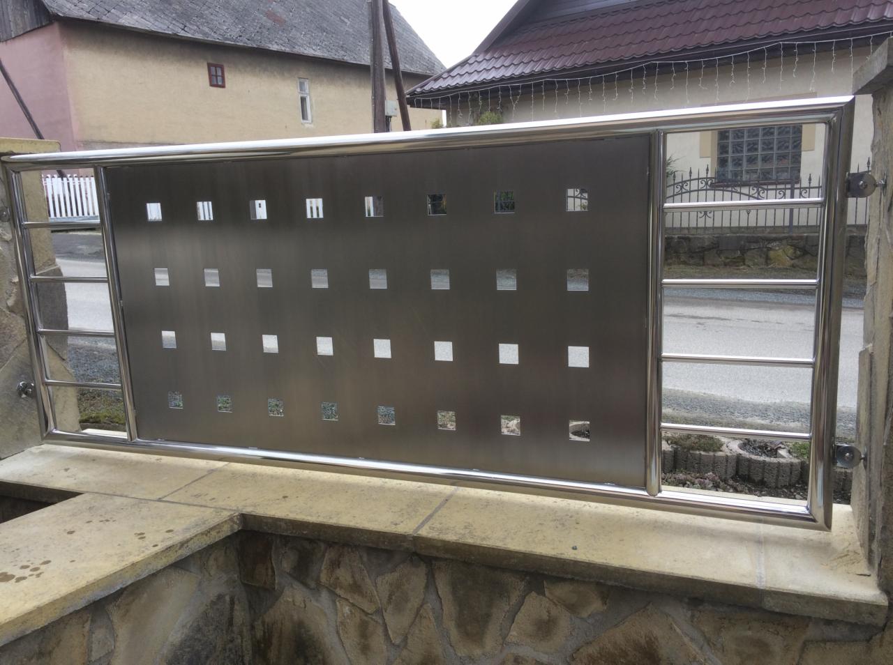 Nowoczesna balustrada ze stali nierdzewnej z perforowanym panelem i kwadratowymi otworami, zamontowana na kamiennym murku przed budynkiem.