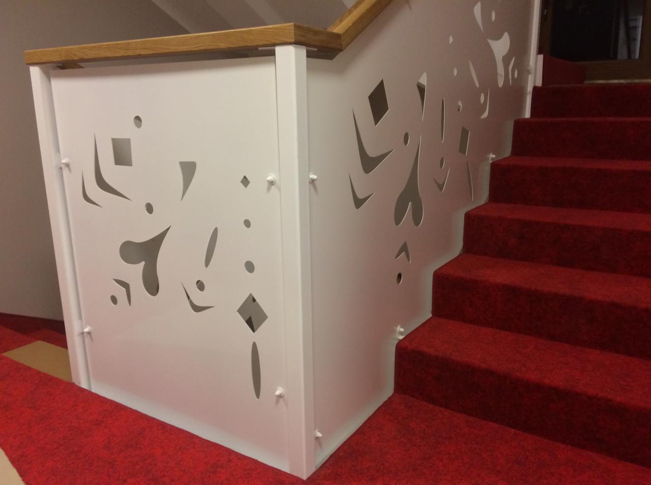 Biała balustrada schodowa z wycinanymi wzorami geometrycznymi i sercami, połączona z drewnianą poręczą i czerwonym dywanem na schodach.
