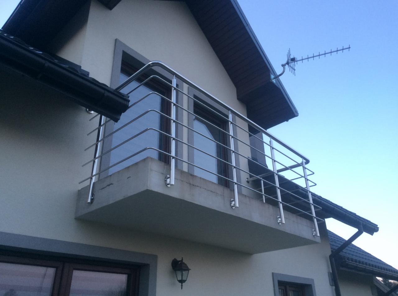 Balkon z prostą, stalową konstrukcją zabezpieczającą, widok z dołu, z anteną na dachu budynku w tle.
