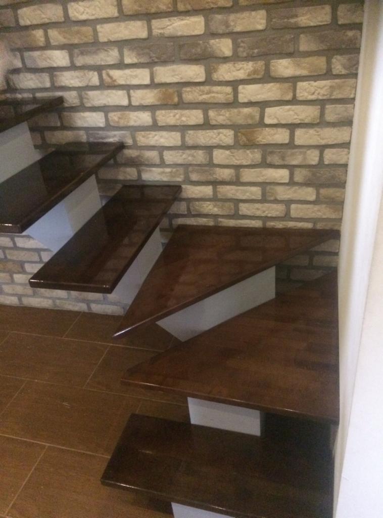 Stopnie schodów z ciemnego drewna, wsparte na białych konstrukcjach, przylegające do ściany z cegły w odcieniach beżu i brązu.