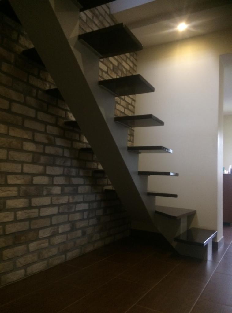 Nowoczesne schody o minimalistycznej konstrukcji z ciemnymi stopniami i szarą belką nośną, przylegające do ściany z cegły dekoracyjnej i białej ściany.