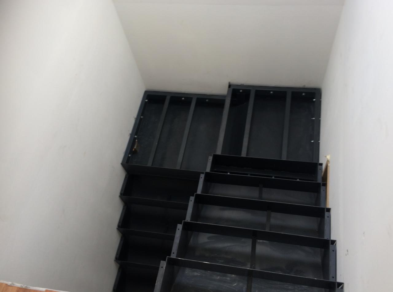 Niedokończona konstrukcja metalowych schodów z czarnych profili, widok z góry, surowe białe ściany i sufit w tle.