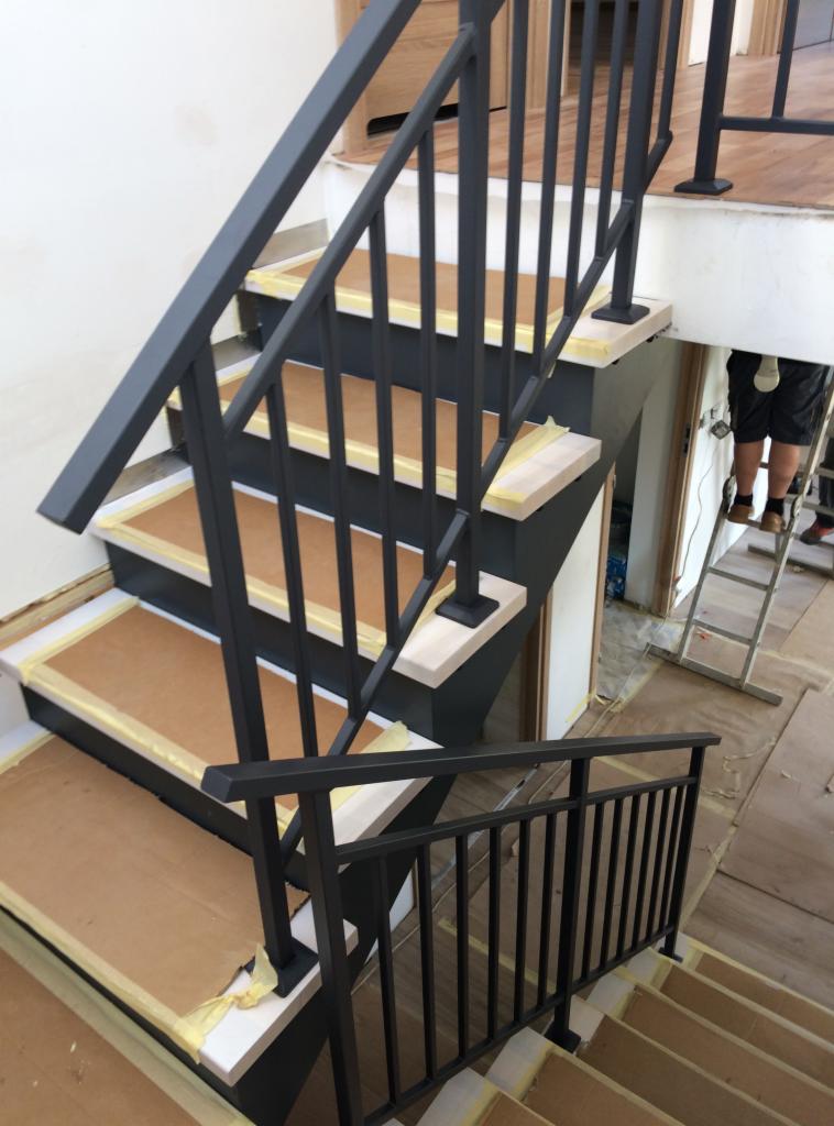 Montaż czarnej, metalowej balustrady schodowej z pionowymi prętami, widoczne zabezpieczenia schodów kartonem i taśmą malarską, drabina w tle.
