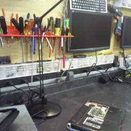 Stanowisko pracy serwisanta komputerowego: otwarty laptop z widocznymi podzespołami, narzędzia na ścianie, dwa monitory, klawiatura i liczne kable na blacie.