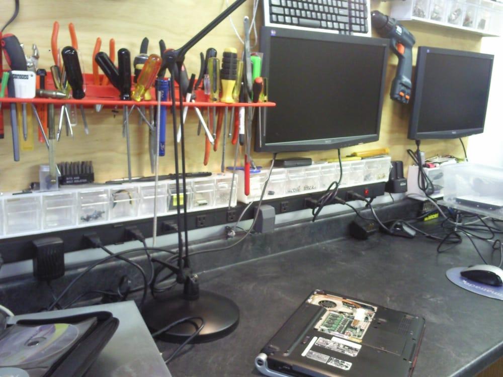 Stanowisko pracy serwisanta komputerowego: otwarty laptop z widocznymi podzespołami, narzędzia na ścianie, dwa monitory, klawiatura i liczne kable na blacie.