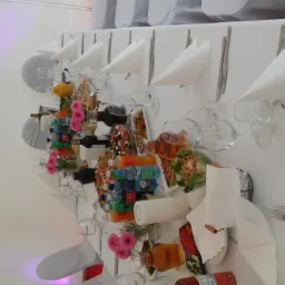 Uroczysty stół w białym namiocie: nakrycia z białymi serwetkami, szkło, kwiatowe dekoracje i napoje, widok z boku.